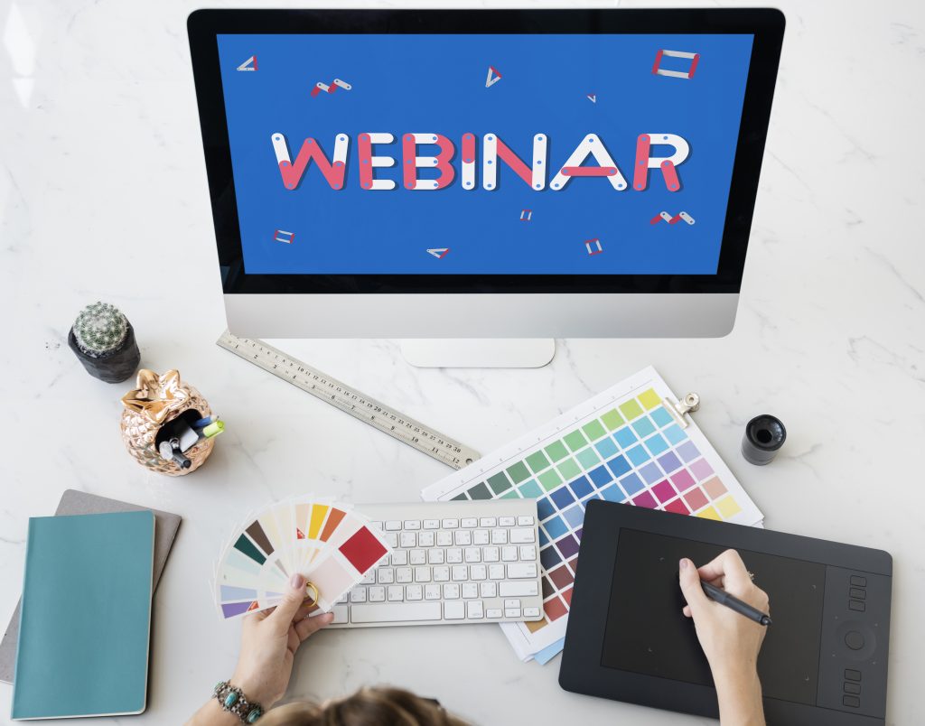 Designer sitzt vor Desktop auf welchem "Webinar" steht. Vor der Person liegen eine Tastatur, Farbpalleten und ein Grafik Tablet.